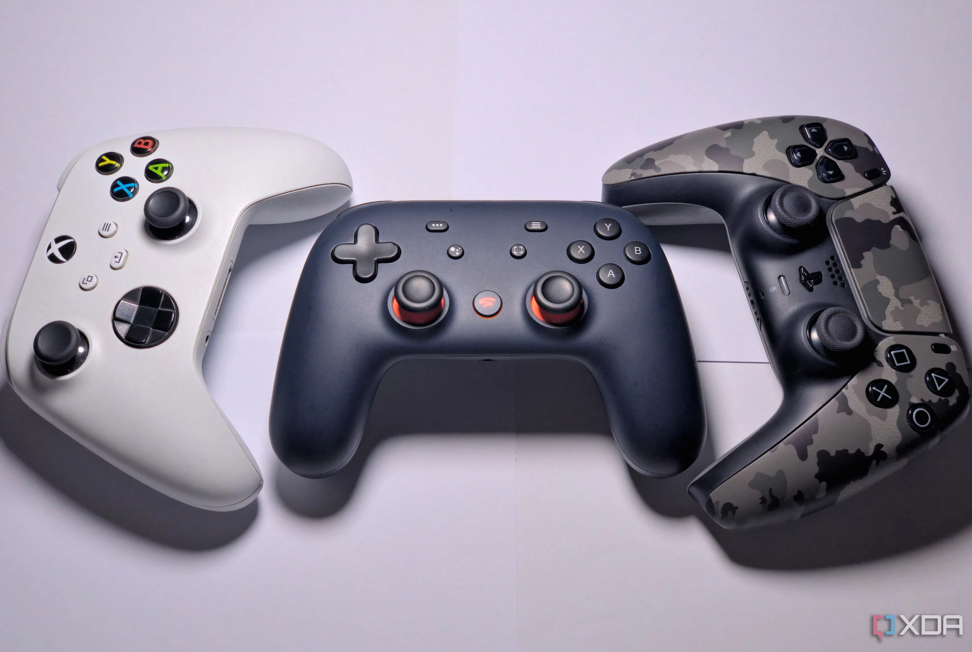 Bộ điều khiển Google Stadia màu xanh đậm đặt cạnh bộ điều khiển Xbox Series màu trắng và bộ điều khiển DualSense camo trên bàn