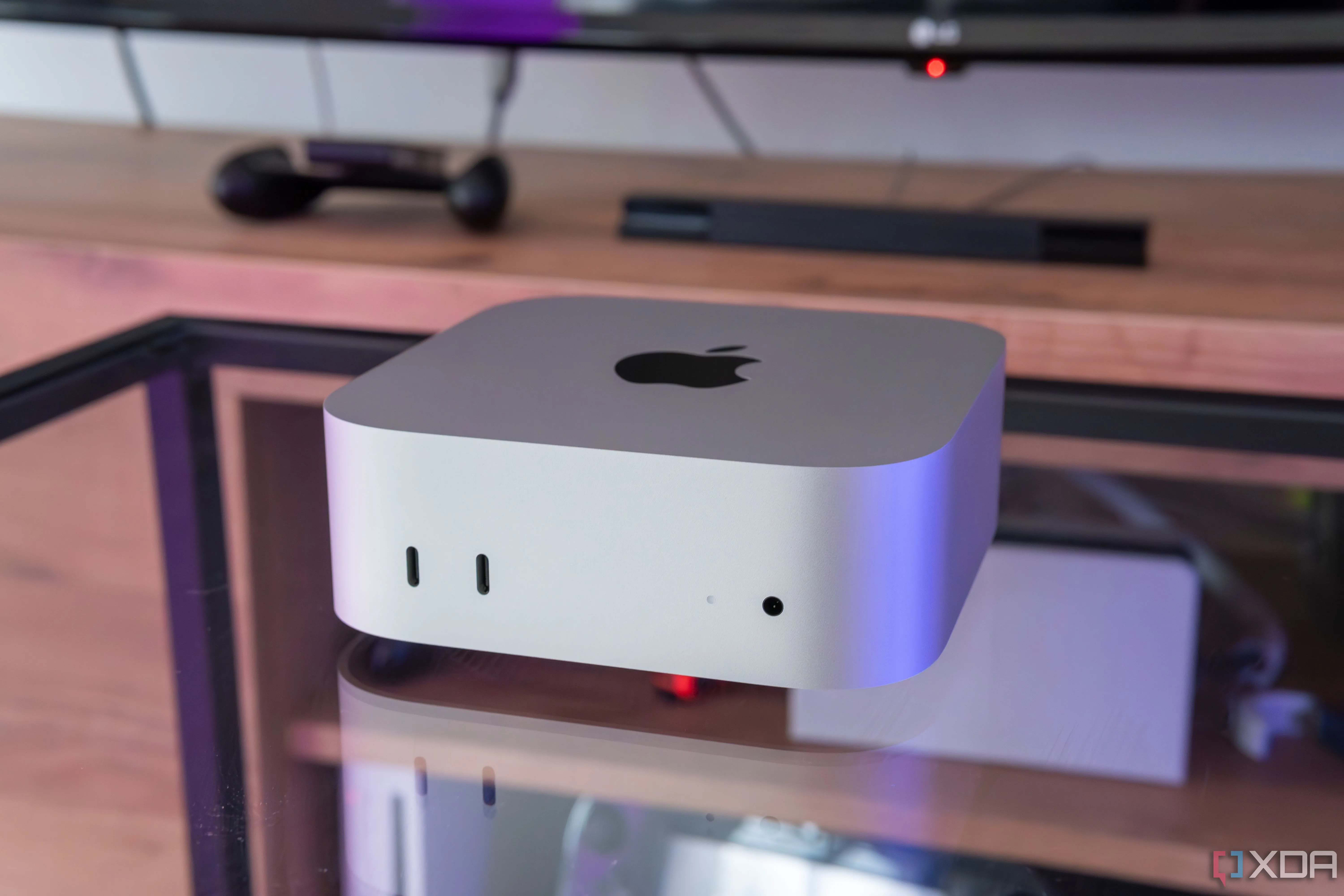 Mac Mini M4 đặt trên bàn kính với kệ TV phía sau, minh họa thiết bị nhỏ gọn của Apple