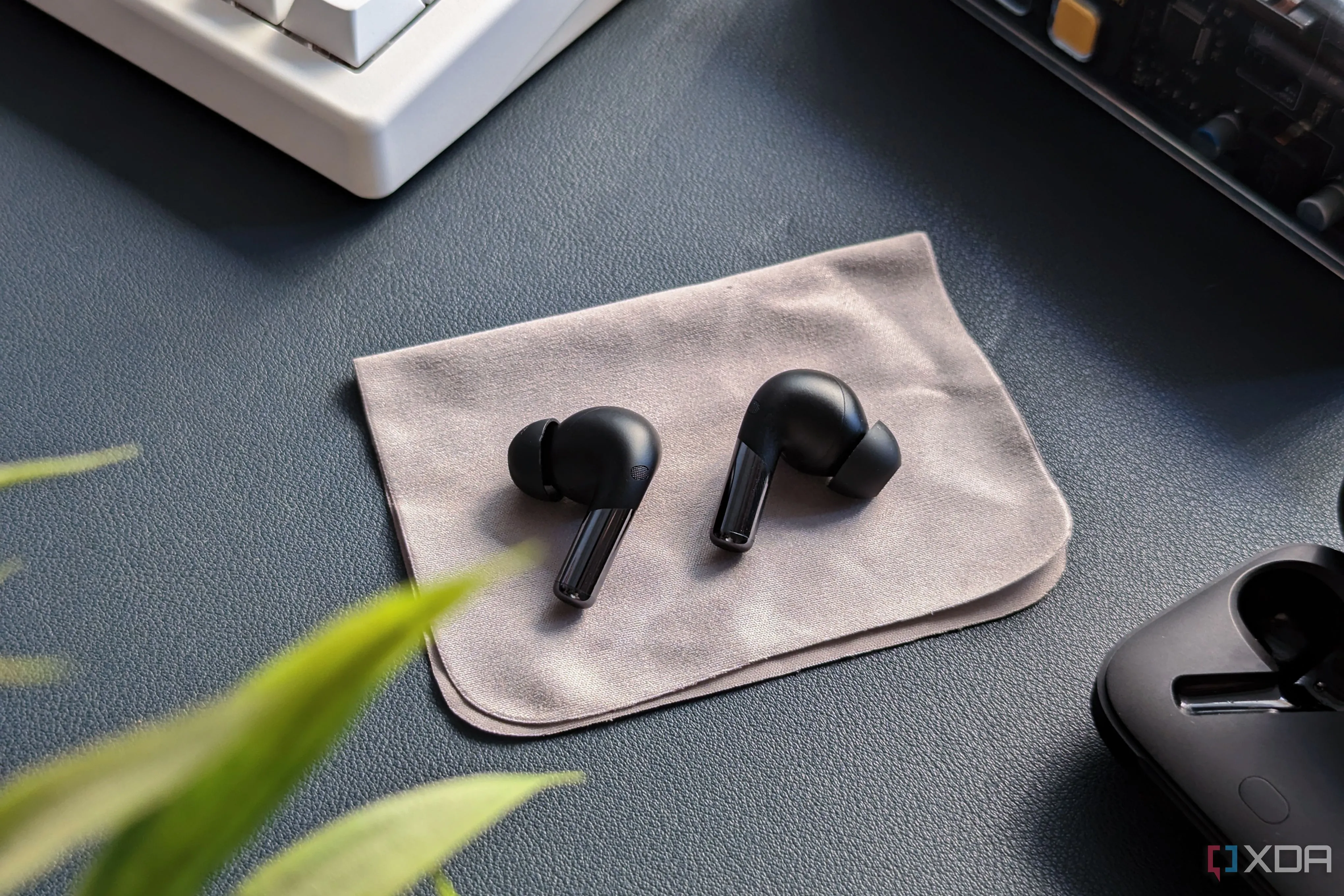 OnePlus Buds Pro 2 placed on microfiber cloth on blue background with its charging case, a keyboard, and a plant in the frame.